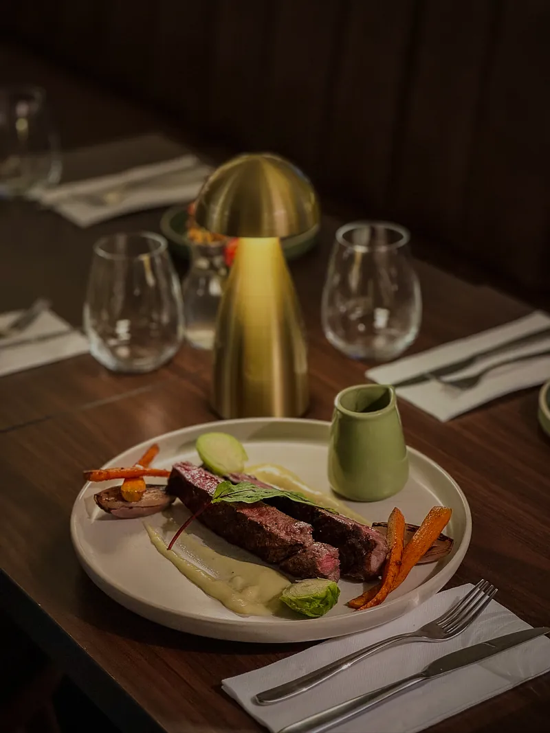 Pièce de bœuf, carottes rôties et purée sur une table dressée, avec une lampe en laiton